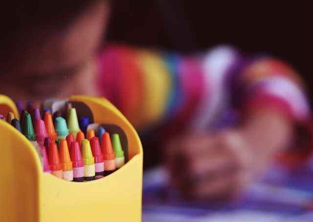 A box of crayons in the foreground as a child does their school work.